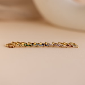Op de afbeelding: Een set van 10 gouden oorringen met een enkele rij gekleurde edelstenen. De oorringen zijn gerangschikt in een regenboogpatroon, beginnend met rood en eindigend met paars.