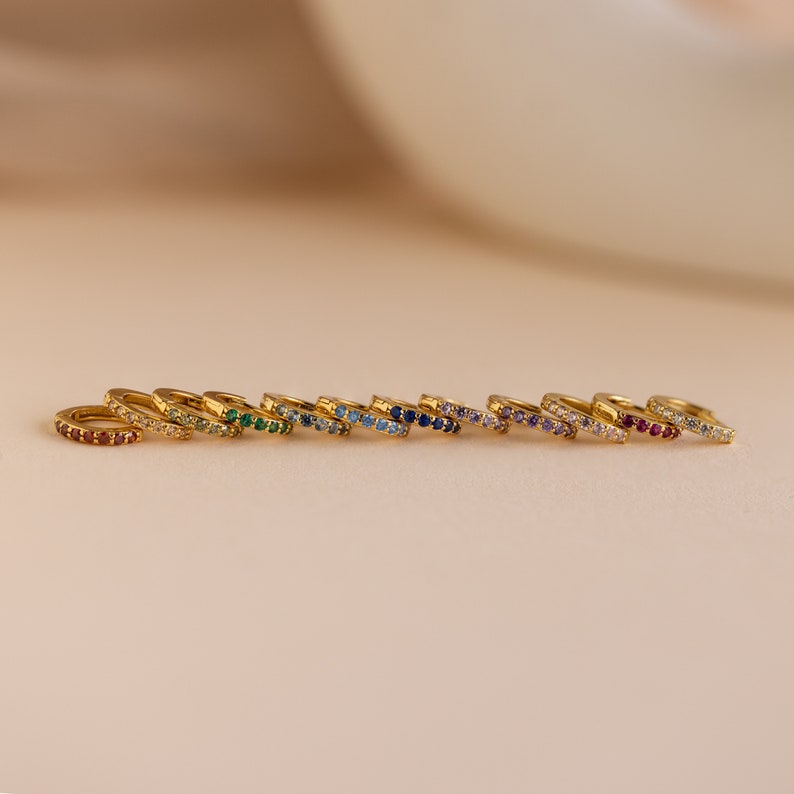 Op de afbeelding: Een set van 10 gouden oorringen met een enkele rij gekleurde edelstenen. De oorringen zijn gerangschikt in een regenboogpatroon, beginnend met rood en eindigend met paars.