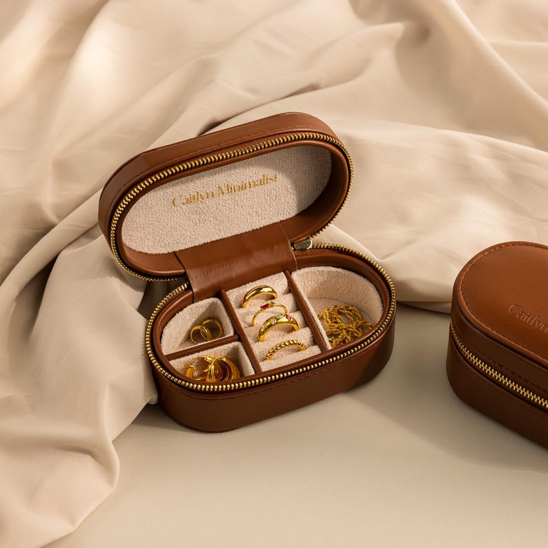 May include: A brown leather oval shaped jewelry box with a zipper closure. The box is open and has multiple compartments for rings, earrings, and necklaces. The box is labeled 'Catherine Michiels'.