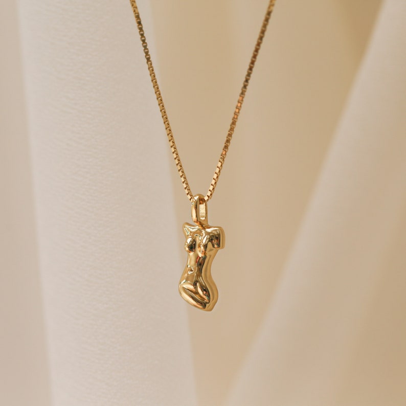 Close-up of a gold chain necklace featuring a sculptural pendant in the shape of a female torso, displayed against a softly lit neutral background.