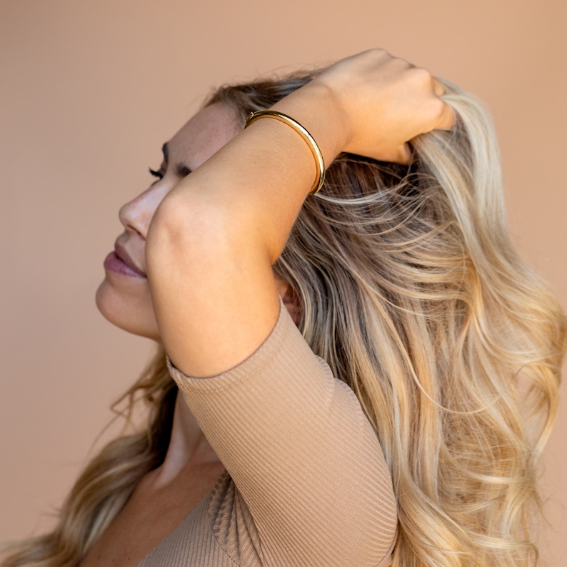 Puede incluir: Una mujer con el pelo largo y rubio lleva una pulsera de oro en la mu&ntilde;eca. Lleva una camisa de manga corta de color marr&oacute;n claro con textura de canal&eacute;.