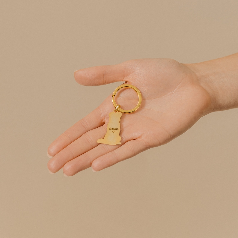 May include: Gold-colored keychain with a dog silhouette and the word "Scout" engraved on it. The keychain is held in a hand.