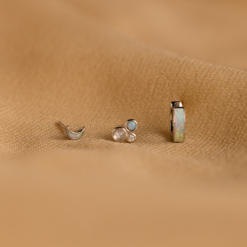 May include: Three silver earrings with opal and diamond accents. The earrings are a crescent moon, a cluster of three circles, and a hoop.
