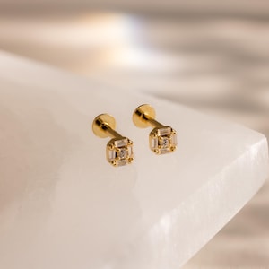 May include: A pair of gold stud earrings featuring a square-shaped head with a central round stone surrounded by four baguette-cut stones. The earrings have a flat back post design, showcasing a polished gold finish and clear, sparkling stones.