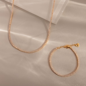 May include: A gold-toned necklace and bracelet set. The necklace is a delicate chain with small, evenly spaced gemstones. The matching bracelet has a similar design, with a heart-shaped charm at the clasp. The jewelry is displayed on a light-colored surface.