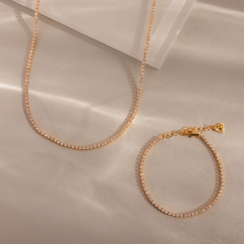 May include: A gold-toned necklace and bracelet set. The necklace is a delicate chain with small, evenly spaced gemstones. The matching bracelet has a similar design, with a heart-shaped charm at the clasp. The jewelry is displayed on a light-colored surface.