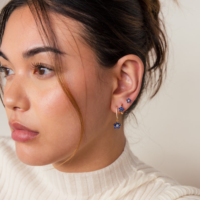 May include: A woman wearing gold hoop earrings with blue flower accents. The earrings are dangling and have a small blue flower charm. She is also wearing a small blue flower stud earring in her upper ear.