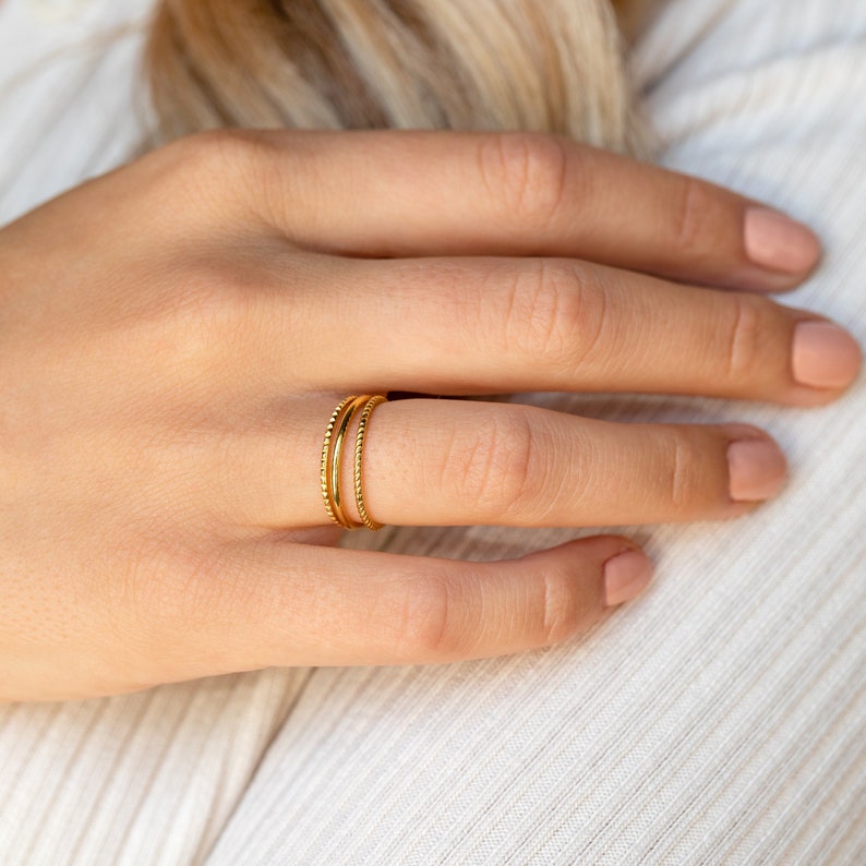A close up of our Dainty Stacking Ring Set in 18K Gold finish placed on on models ring finger - featuring a set of 3 rings: 1 Thin Band, 1 Twist Ring and 1 Notched Ring all about 1mm thick.