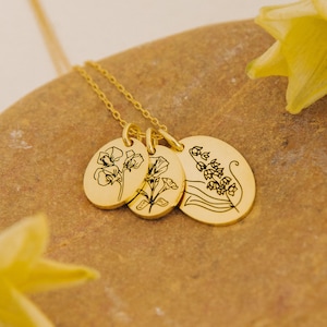May include: A gold-colored necklace with three oval pendants. Each pendant features a black floral design. The necklace is displayed on a textured, light brown surface, with a yellow flower in the background.
