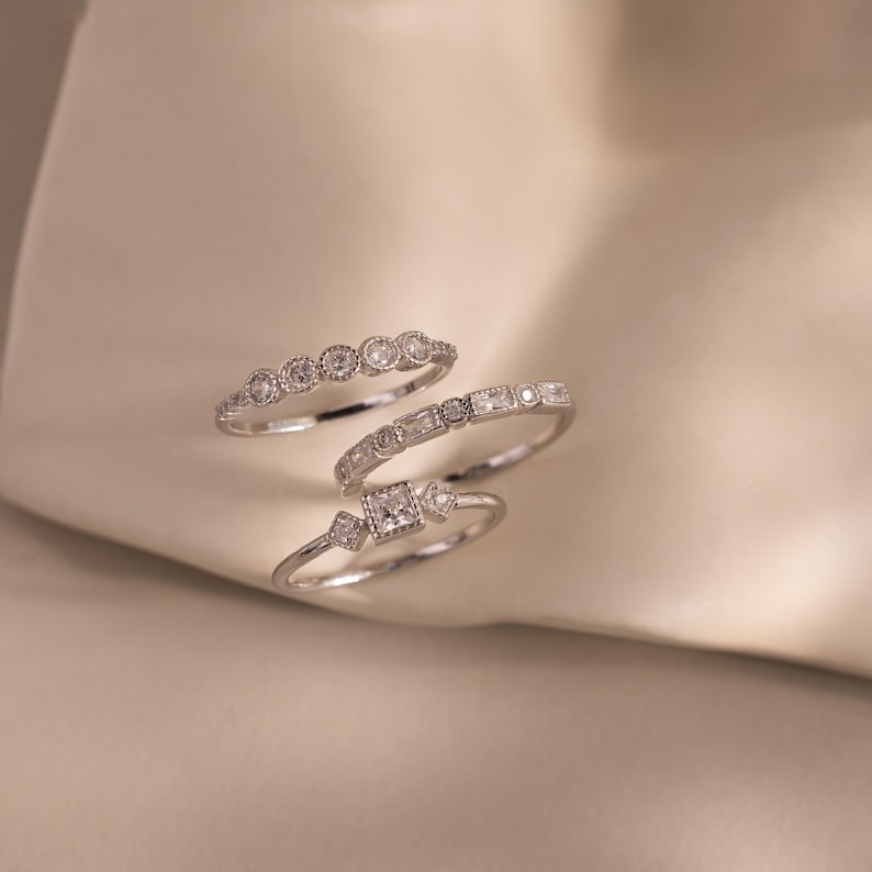 May include: Four silver rings with clear gemstones. The rings feature various designs, including round, baguette, and square-cut stones. The rings are displayed on a soft, off-white surface, creating a delicate and elegant aesthetic.