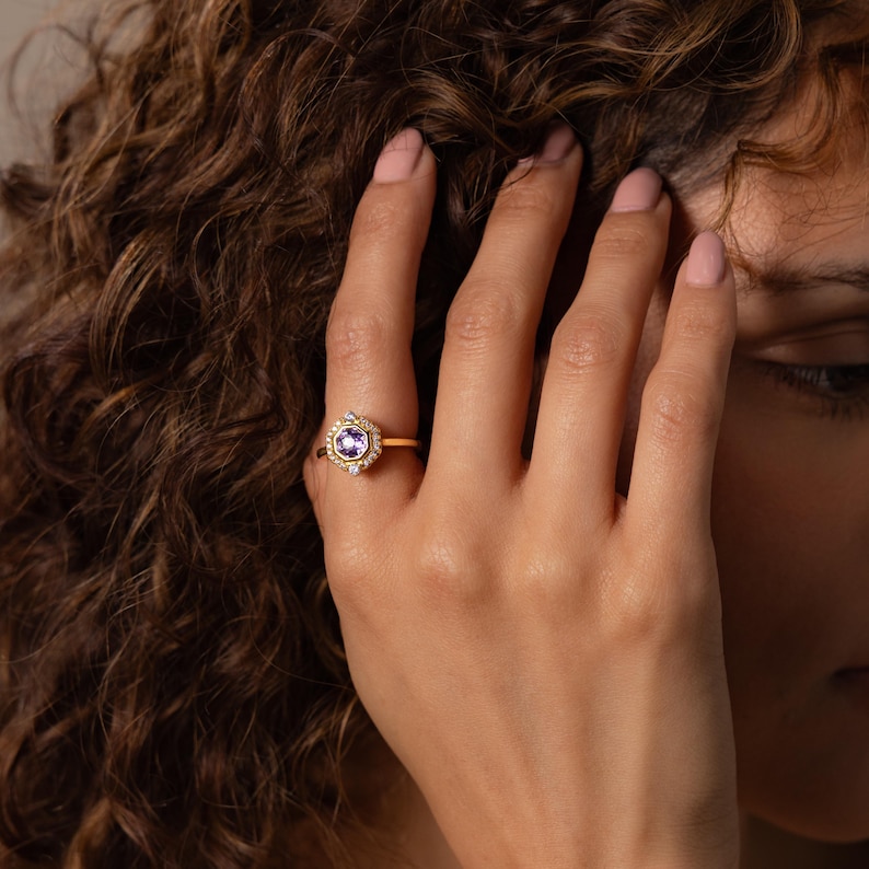 May include: A gold ring featuring a central hexagonal setting with a round purple gemstone. The setting is surrounded by a halo of small clear stones, with additional clear stones accenting the band on either side of the central hexagonal frame.