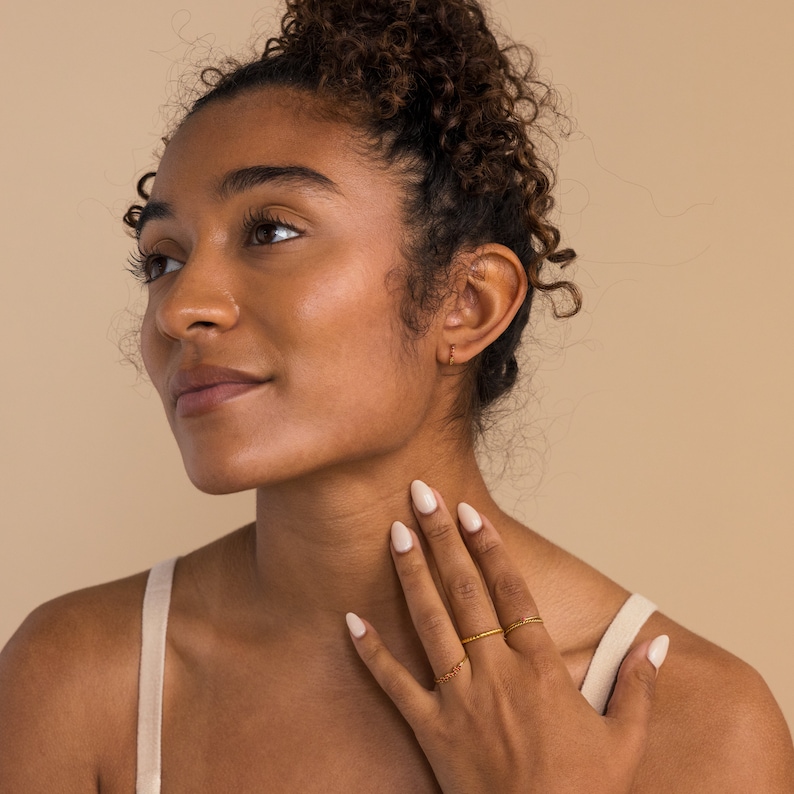 Op de afbeelding: Een vrouw met bruin krullend haar draagt een wit tanktopje en heeft haar hand op haar nek. Ze draagt ​​meerdere gouden ringen aan haar vingers en heeft witte nagellak op haar nagels.