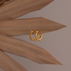 May include: A pair of small gold hoop earrings featuring a textured surface with evenly spaced raised spherical beads along the outer edge. The metallic gold finish reflects light, highlighting the circular beaded pattern on each hoop earring.