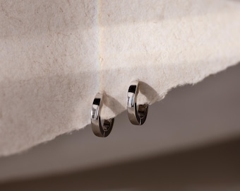 Op de afbeelding: Een paar kleine, gepolijste zilverkleurige oorringen met een glad, reflecterend oppervlak. De oorbellen zijn cirkelvormig en hebben een scharniersluiting, wat zorgt voor een strakke, metalen afwerking.