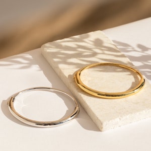 Puede incluir: Dos pulseras simples, una plateada y otra dorada, sobre una superficie blanca. Las pulseras son redondas y tienen un acabado liso y pulido.