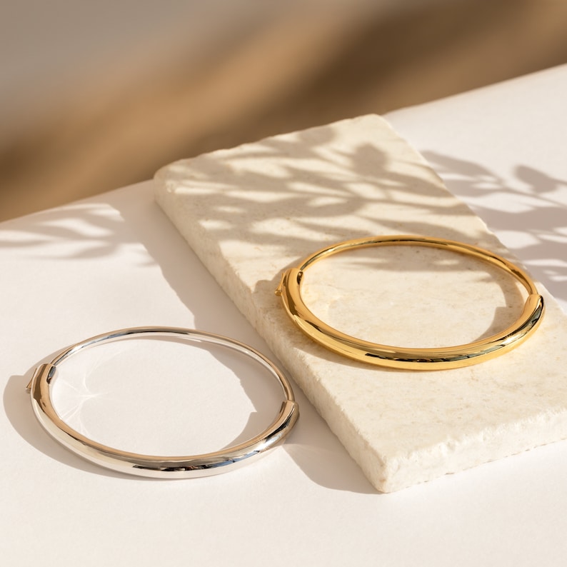 Puede incluir: Dos pulseras simples, una plateada y otra dorada, sobre una superficie blanca. Las pulseras son redondas y tienen un acabado liso y pulido.