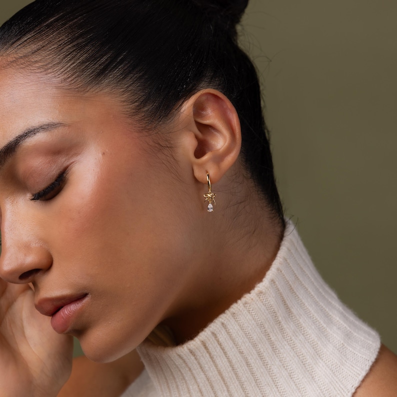 May include: A gold-colored hoop earring with a bow design and a small, dangling, clear gemstone. The earring is worn on the ear of a person wearing a white sweater. The background is a solid, muted green.