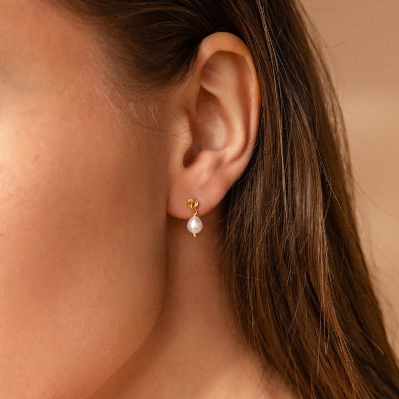 May include: A close-up of a gold-colored earring with a small, white pearl. The earring is attached to a person's ear. The earring has a small loop design at the top.