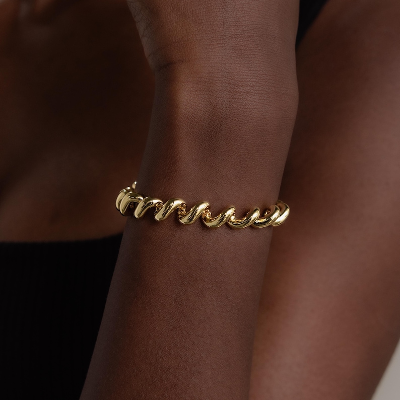 May include: A gold bracelet with a spiral design. The bracelet is worn on an arm and is made of a shiny, metallic material. The bracelet's design is a continuous, twisted rope-like pattern.