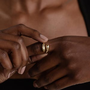 May include: A gold ring featuring an open design with two smooth, polished, teardrop-shaped ends that face each other. The metal has a reflective, high-shine finish, creating a fluid and organic appearance on the finger with a gap between the ends.