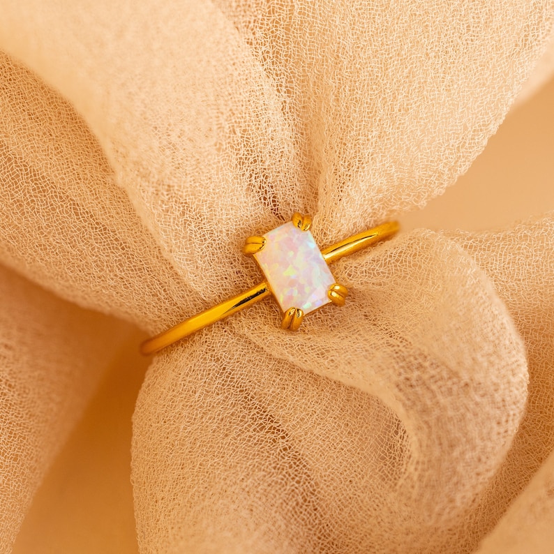 May include: A gold ring with a rectangular opal gemstone. The opal has a white and iridescent appearance. The ring is set on a light beige fabric.