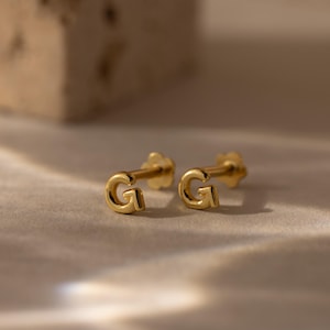 Pair of gold letter G stud earrings with a smooth rounded finish, displayed on a softly lit stone surface with warm neutral tones.