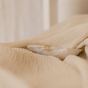 May include: A pair of gold earrings with small, clear stones set in a delicate floral design. The earrings are displayed on a white, textured surface.