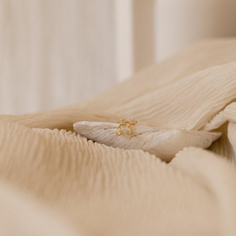 May include: A pair of gold earrings with small, clear stones set in a delicate floral design. The earrings are displayed on a white, textured surface.