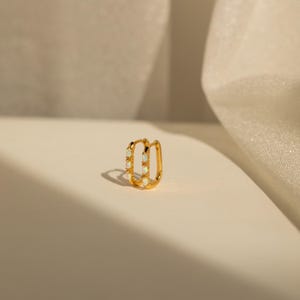 May include: A gold-toned rectangular hoop earring featuring three small, vertically aligned oval-shaped white opals set along the front edge. The earring has a polished metallic finish and a hinged closure mechanism visible at the top.
