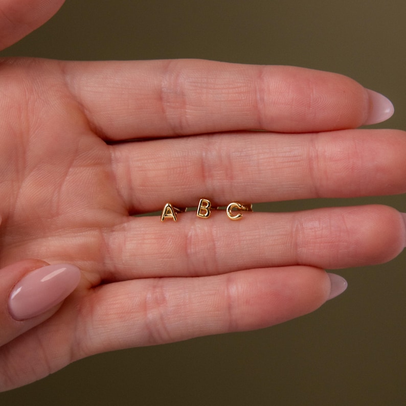 Three tiny gold initial stud earrings with the letters A, B, and C resting across a hand, showing their small scale against the fingers.