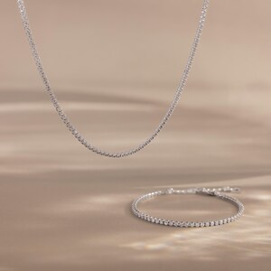 May include: A delicate silver necklace and matching bracelet, both adorned with small, sparkling stones. The necklace hangs gracefully, while the bracelet is positioned below, creating a coordinated jewelry set. The background is a soft, neutral tone.