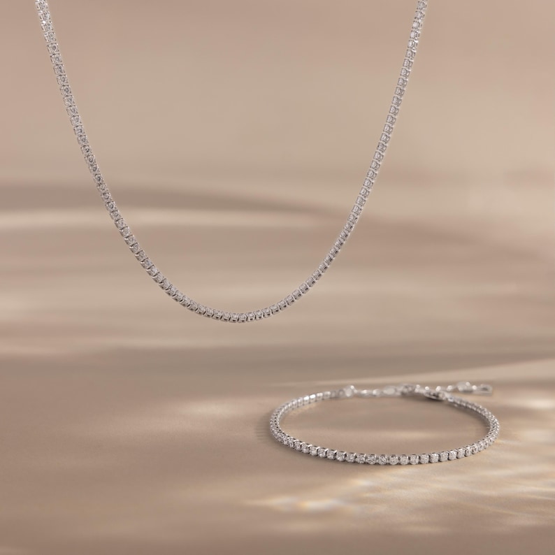 May include: A delicate silver necklace and matching bracelet, both adorned with small, sparkling stones. The necklace hangs gracefully, while the bracelet is positioned below, creating a coordinated jewelry set. The background is a soft, neutral tone.