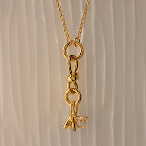 May include: A gold-colored necklace with a chain and a pendant featuring the letters "A" and "Q". The pendant is connected to a series of interlocking rings and a small charm. The necklace is displayed against a white, textured background.
