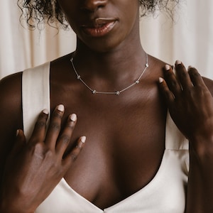 May include: A close-up of a person wearing a white tank top with a delicate silver necklace featuring small star-shaped charms.