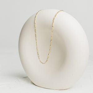 May include: A delicate gold-colored Figaro chain necklace displayed on a white, rounded pedestal. The necklace features a repeating pattern of elongated and circular links, creating a subtle, elegant design. The chain is presented against a clean, minimalist backdrop.