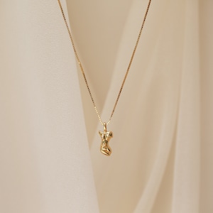 May include: A gold necklace with a pendant shaped like a stylized human figure. The pendant is textured and has a slightly abstract design.