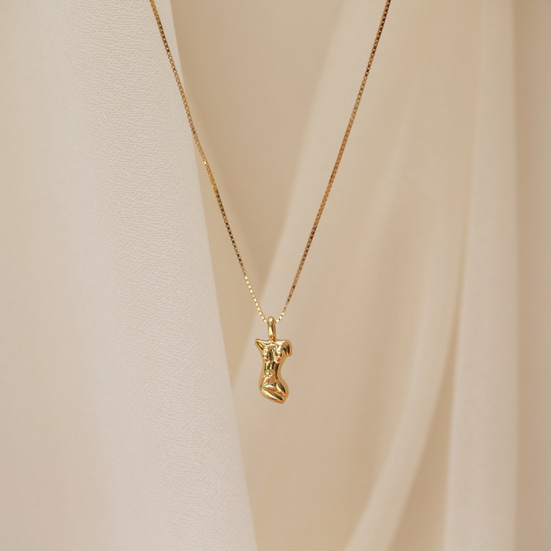 May include: A gold necklace with a pendant shaped like a stylized human figure. The pendant is textured and has a slightly abstract design.