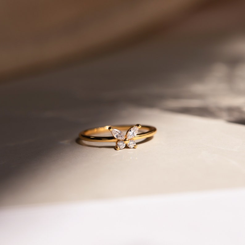 A close up of our Mariposa Butterfly Ring in 18K Gold finish placed on a white table with natural light coming in from the right side - featuring an about 5.5mm x 8mm CZ Diamond Butterfly Charm and an about 1.5mm Thin Ring Band.