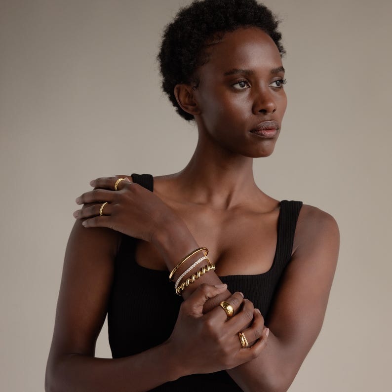 May include: A selection of gold jewellery, including rings and bracelets, is displayed on a person's hands and arms. The bracelets include a thick gold band, a twisted rope design, and a band with small, clear stones. The rings are various styles.