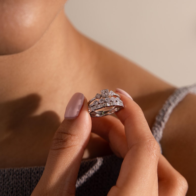 May include: A set of three silver rings with multiple clear gemstones. The rings are held in a person's hand. The top ring has a square gemstone surrounded by smaller stones. The middle ring has a row of small stones. The bottom ring is a simple band.