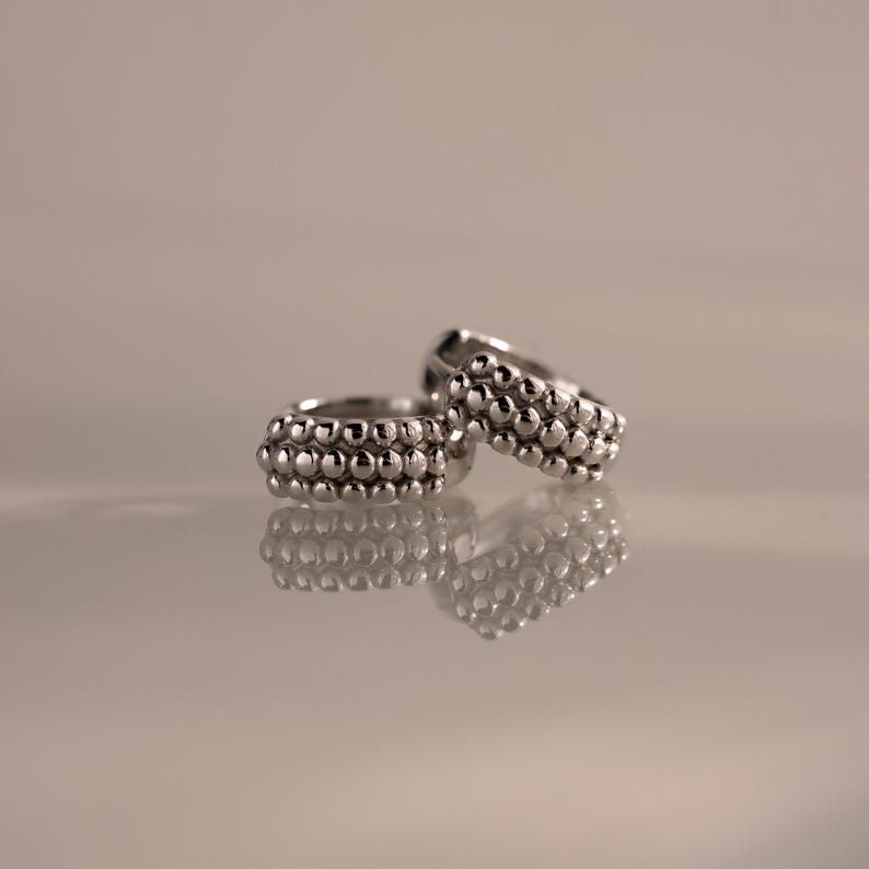 May include: Two silver rings with a textured design of small, round beads. The rings are displayed on a reflective surface, showcasing their intricate details and polished finish. The rings are a jewelry item.