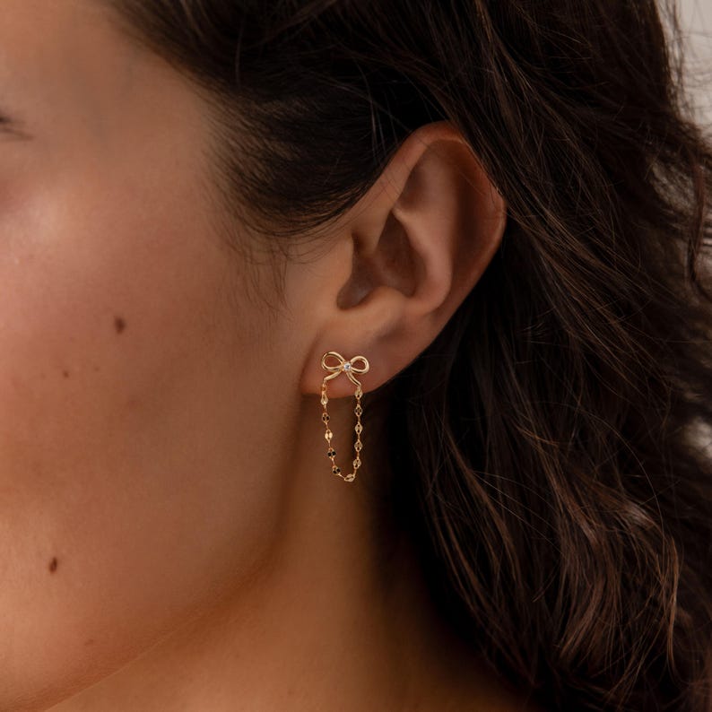 Woman wearing Bow Chain Stud Earrings in 18K Gold. It features a bow stud with a diamond in the middle, with a mirror chain dropping  in a loop shape from the bow
