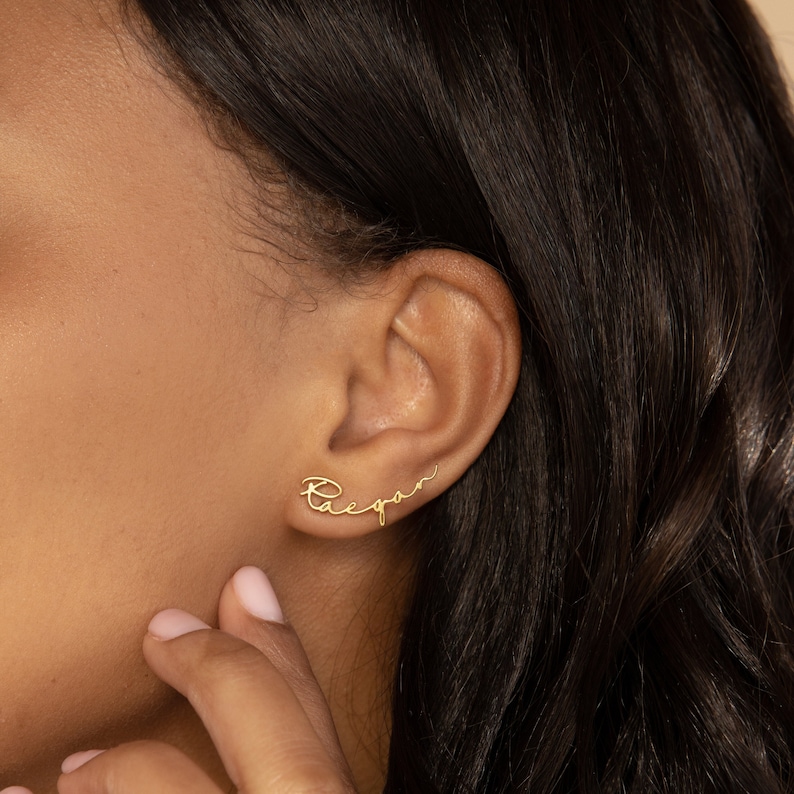 Dainty gold stud name earrings sit it in the first piercing of the ear. The charm earring has the name 'Raegan' spelled in a minimalist script font.