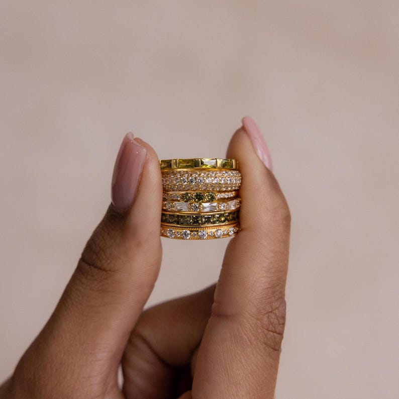 May include: A stack of gold-toned rings with various designs, including bands with clear and green gemstones. The rings are held in a hand with manicured nails, set against a neutral background. The rings are likely for fashion or jewelry.