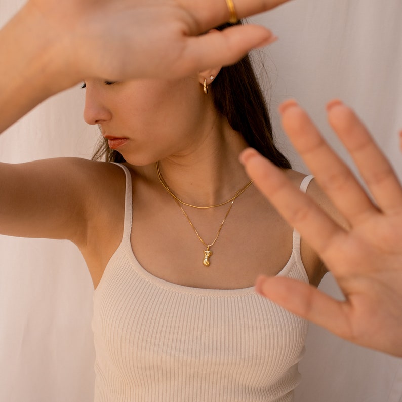May include: A woman wearing a white tank top with a gold chain necklace and a gold pendant. She is wearing gold hoop earrings and a gold ring on her finger.