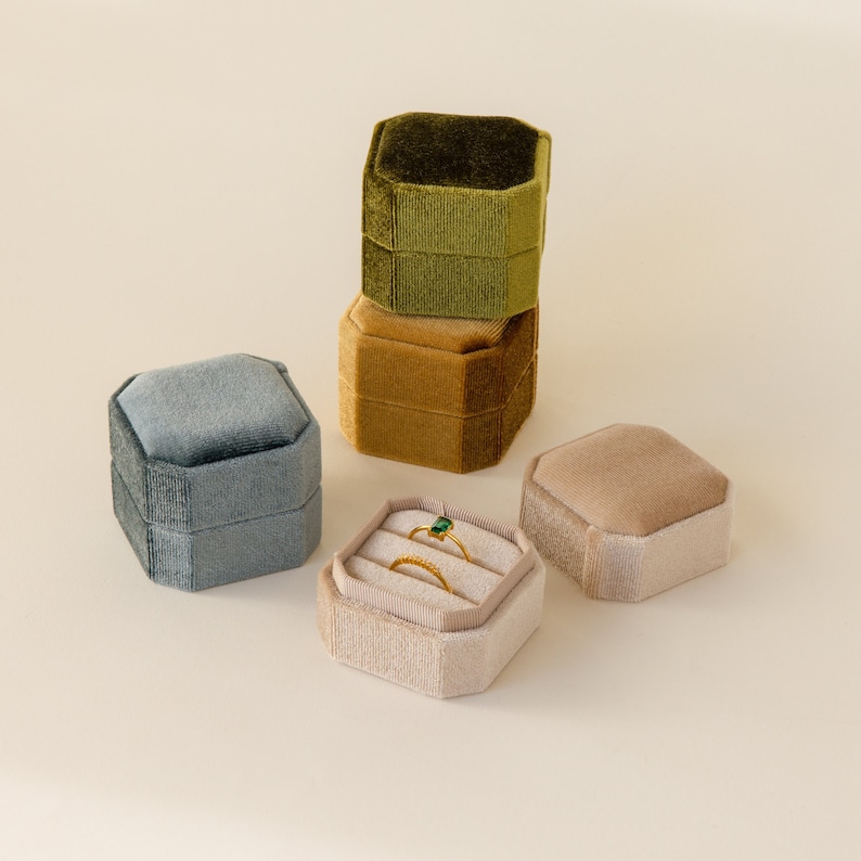 May include: Four velvet ring boxes in different colors. The boxes are stacked on top of each other. The top box is green, the second box is gold, the third box is blue, and the bottom box is beige. The beige box is open and contains two rings.
