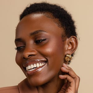 May include: A pair of gold-toned earrings featuring a knotted, multi-layered design. The earrings have a polished, reflective surface and a rounded, intertwined structure that creates a compact, circular shape worn on the earlobe.