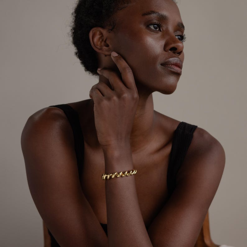 May include: A gold bracelet featuring a twisted rope design. The bracelet is worn on a wrist. The person is wearing a black vest top. The background is a neutral beige colour.
