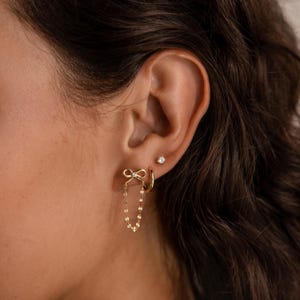Woman wearing Bow Chain Stud Earrings in gold with delicate bow studs and draped chain loops, styled in a stacked ear look with a small stud and hoop.