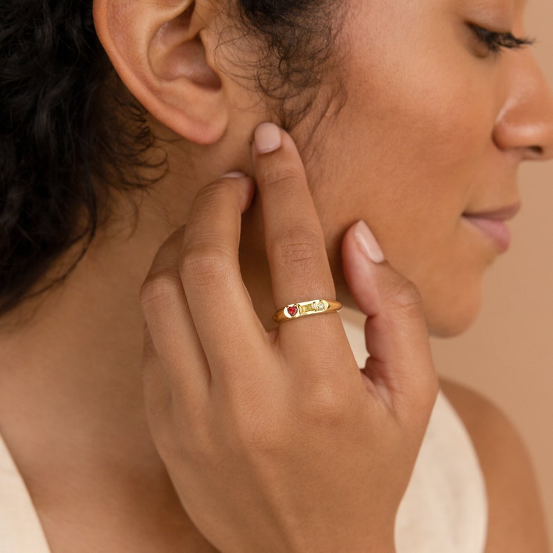 A gold signet ring with birthstones is on the pointer finger. There is a Ruby heart stone, a Topaz baguette stone, and a clear crystal circle stone next to each other.